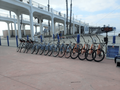 A row of colorful bicycles lined up along a beachfront walkway near a pier.