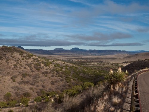 A scenic view of rolling hills and mountains under a blue sky, with a winding road in the foreground.