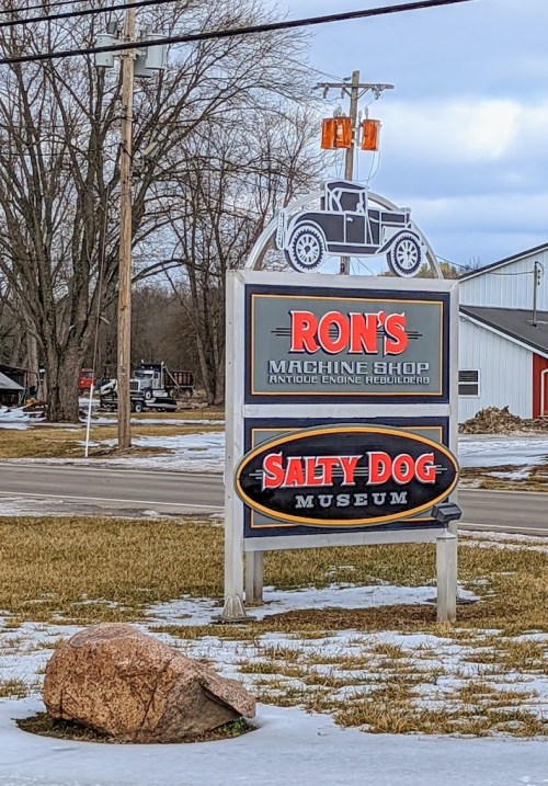 Sign for Ron's Machine Shop and Salty Dog Museum, featuring a vintage car silhouette and surrounded by snow.
