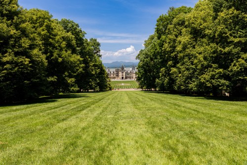 A lush green lawn leads to a grand mansion, framed by tall trees and mountains in the background under a blue sky.