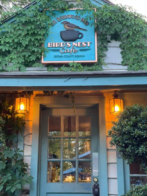 A charming café entrance with a sign reading "Bird's Nest Café," surrounded by green ivy and warm outdoor lighting.