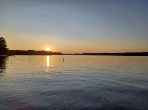 A serene lake at sunset, with calm waters reflecting the orange and blue sky. Trees line the distant shore.