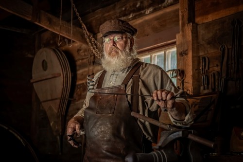 A bearded blacksmith in a workshop, wearing glasses and a cap, holding a tool with various tools and equipment in the background.