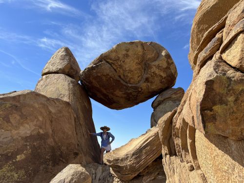 A person stands beneath a large boulder balanced on top of two rock formations against a blue sky.