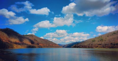 A serene lake surrounded by hills under a bright blue sky with fluffy clouds.