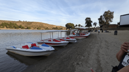A row of colorful boats lined up on a sandy shore by a calm lake, with trees and hills in the background.