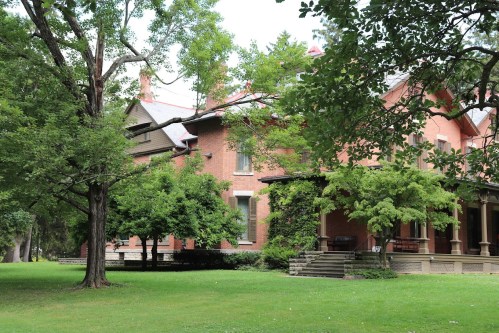 A large brick house surrounded by lush green trees and grass, featuring a porch and multiple windows.