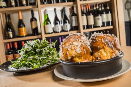 Two golden, cheesy bread rolls in a dish, accompanied by a fresh green salad on a black plate, with wine bottles in the background.