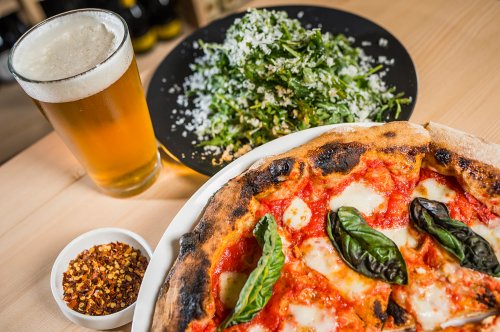 A close-up of a pizza with basil, a salad, a glass of beer, and a bowl of red pepper flakes on a wooden table.