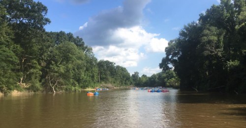 A serene river scene with people floating on colorful tubes, surrounded by lush green trees and a blue sky with clouds.