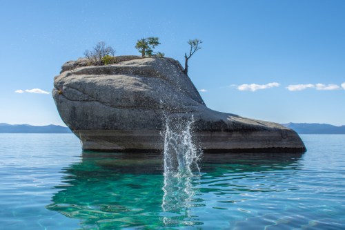A large rock formation with a small tree on top, surrounded by clear blue water and a splash from a diving point.