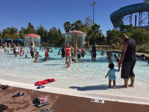 A busy water park pool with families and children playing, surrounded by palm trees and water features under a clear blue sky.
