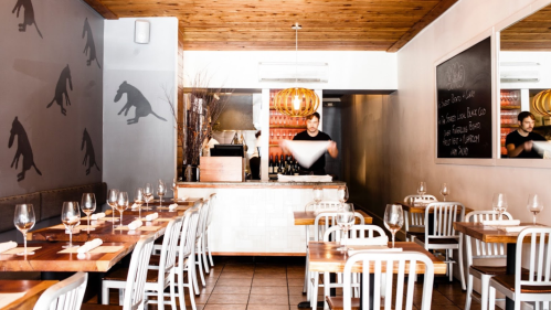 A modern restaurant interior with wooden tables, white chairs, and a bartender preparing drinks in the background.