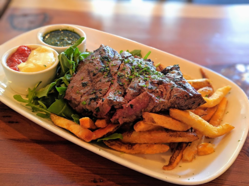 A plate of grilled steak on a bed of fries, served with dipping sauces and a side of greens.