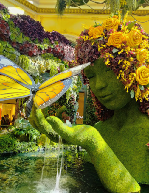 A floral sculpture of a woman with a butterfly on her hand, surrounded by vibrant greenery and flowers.