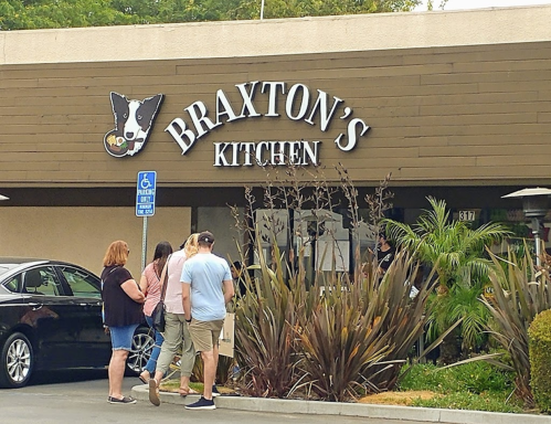 A group of people stands outside Braxton's Kitchen, a restaurant with a sign featuring a dog logo.