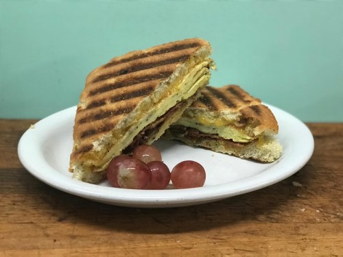 A toasted sandwich with layers of egg and meat, served on a plate with a few grapes.