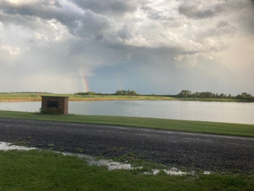 A serene landscape featuring a lake, green grass, and a rainbow peeking through stormy clouds in the background.