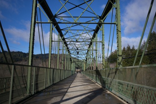 A green metal truss bridge spans a wide path, surrounded by trees and a blue sky with scattered clouds.