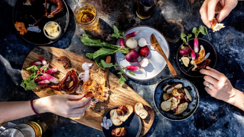 A table spread with various dishes, including meats, vegetables, and drinks, with hands reaching for food.