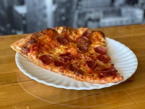 A slice of pepperoni pizza on a white plate, resting on a wooden table with a blurred cityscape background.