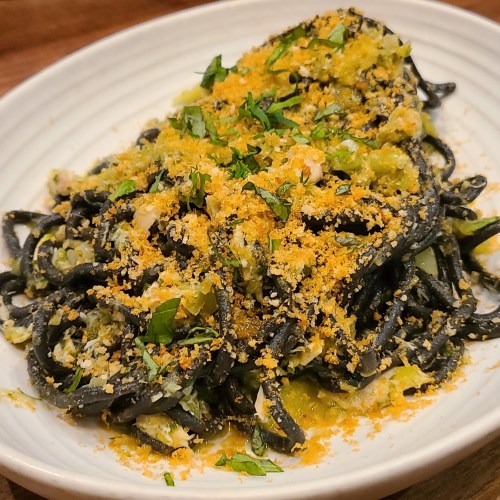 A plate of black spaghetti topped with herbs, breadcrumbs, and green sauce, served on a white dish.