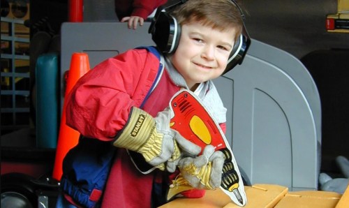 A young boy wearing headphones and gloves smiles while holding a toy saw, engaged in a hands-on activity.