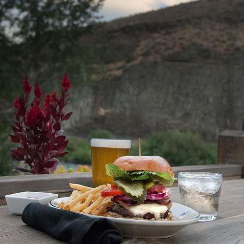 A gourmet burger with fries, a glass of beer, and a glass of water on a wooden table with a scenic background.