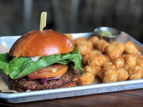 A juicy burger with lettuce, tomato, and onion, served with crispy tater tots on a metal tray.