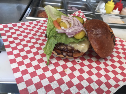 A grilled burger with melted cheese, lettuce, tomatoes, pickles, and onions on a red and white checkered paper.
