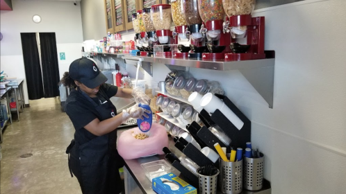 A worker in a black uniform prepares cotton candy at a counter with various cereal dispensers and utensils.