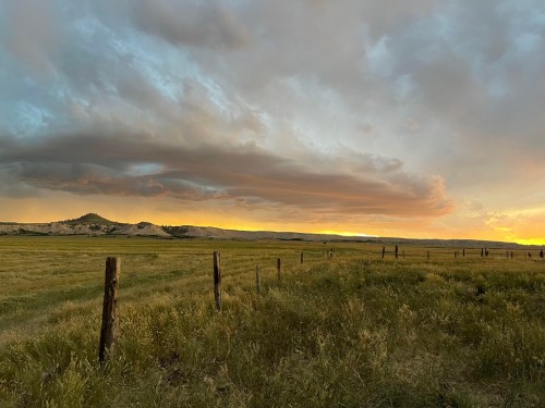 A serene landscape at sunset, featuring rolling hills, a grassy field, and dramatic clouds in the sky.