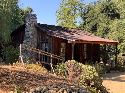 A rustic wooden cabin with a stone chimney, surrounded by greenery and a gravel path.