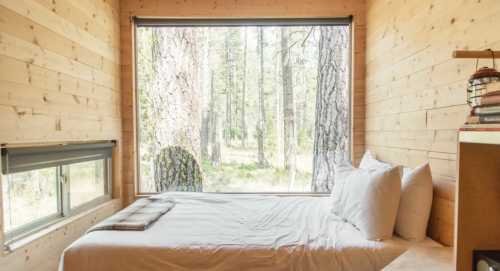 Cozy bedroom with a large window overlooking a forest, featuring a bed with white linens and wooden walls.