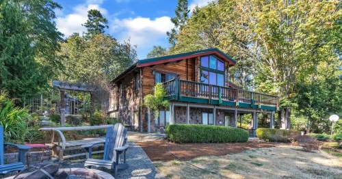 A charming wooden house with a balcony, surrounded by trees and a landscaped yard, under a blue sky.