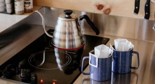 A stainless steel kettle on a stovetop next to two blue mugs with coffee filters inside.