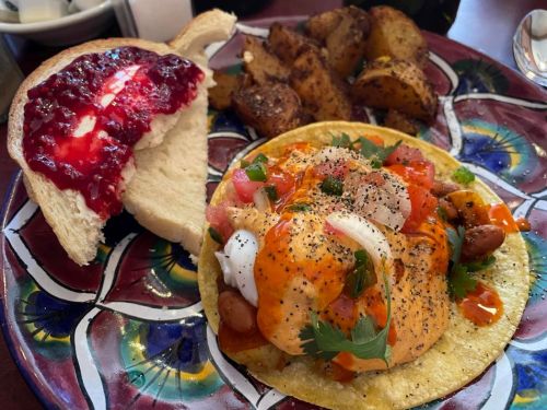 A colorful plate featuring a taco with beans, tomatoes, and sauce, alongside a slice of bread with jelly and roasted potatoes.