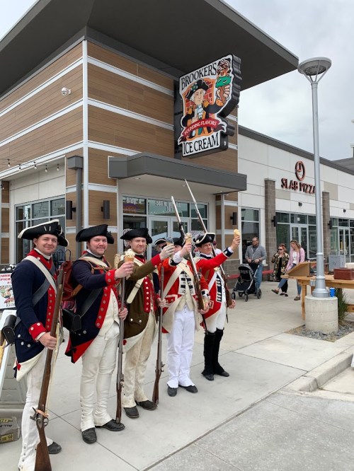 Five men in Revolutionary War uniforms stand outside an ice cream shop, holding ice cream cones and muskets.