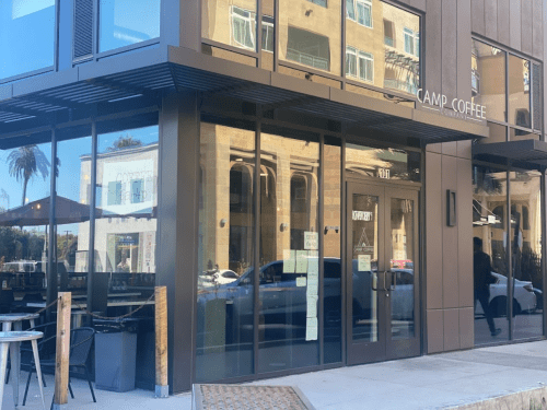 Exterior view of Camp Coffee with large glass windows reflecting nearby buildings and palm trees.