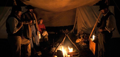 Four people in period clothing gather around a campfire under a tent, sharing stories in a dimly lit setting.