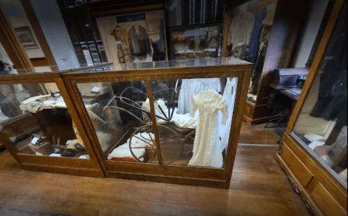 Display case in a museum featuring historical clothing and artifacts, with wooden flooring and surrounding exhibits.