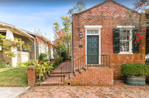 A charming brick house with a green door, surrounded by greenery and autumn foliage.