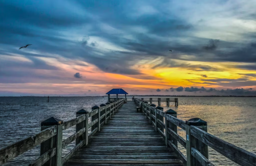 A wooden pier extends into a calm sea under a colorful sunset with clouds and a hint of a bird in the sky.