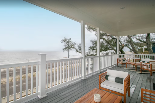 A spacious porch with wooden furniture overlooks a calm beach and ocean under a clear sky.
