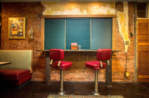 Two red bar stools at a small counter in a rustic brick-walled diner with blue blinds and peeling paint.