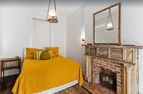 Cozy bedroom with a yellow bedspread, vintage fireplace, and a large mirror on the wall. Bright and inviting atmosphere.