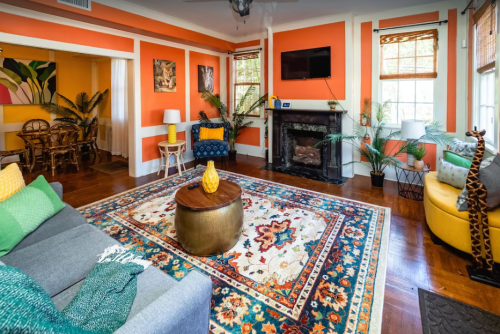 Cozy living room with orange walls, a patterned rug, a fireplace, and tropical decor, featuring a mix of seating options.