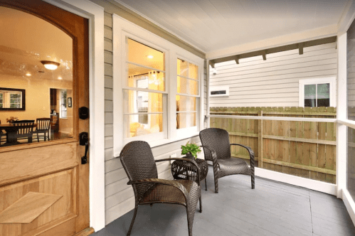 A cozy porch with two wicker chairs and a small plant, featuring a wooden door and a view of a fenced yard.