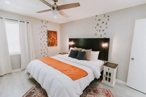 Cozy bedroom with a large bed, orange throw, decorative pillows, and modern lighting against a neutral wall.