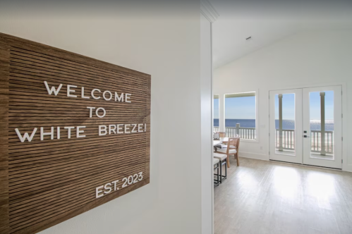 A wooden sign reading "Welcome to White Breeze! EST. 2023" in a bright, airy room with ocean views.